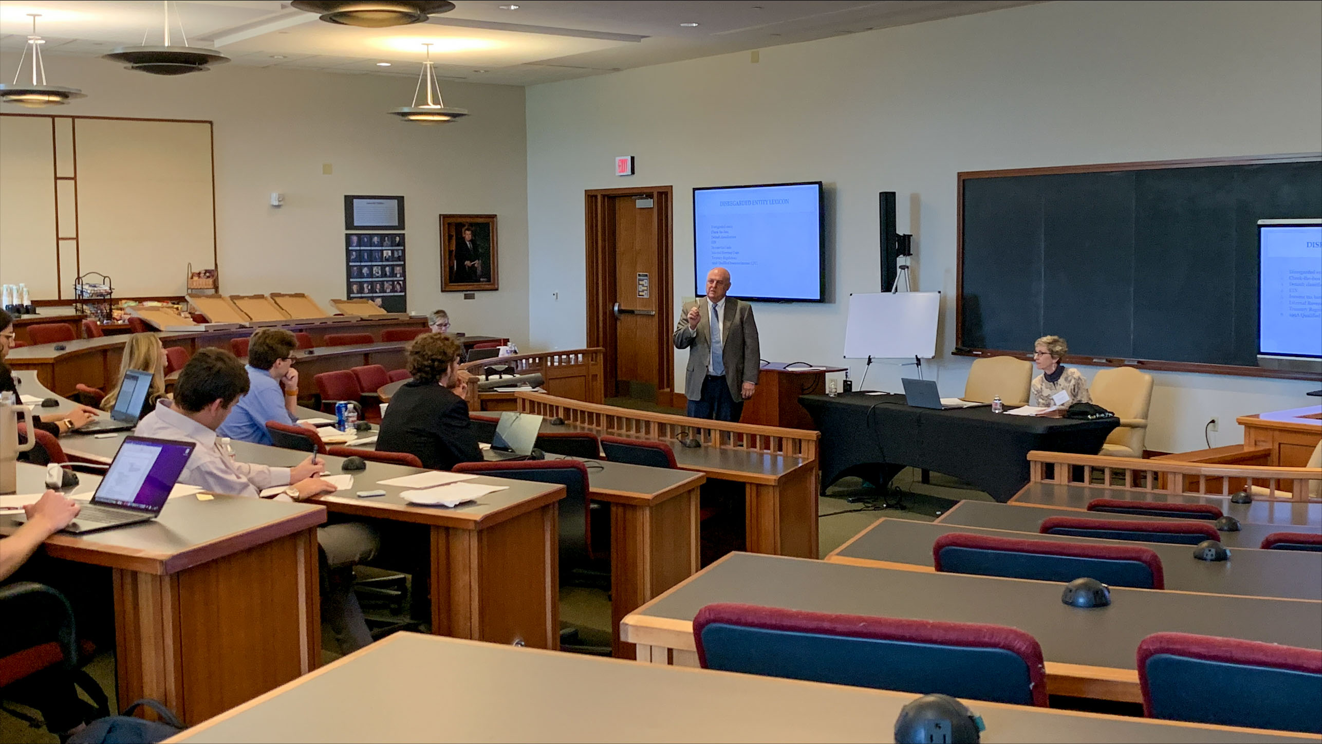 Dan Baucum, standing, and Professor Miller, seated, teach in front of classroom full of students.