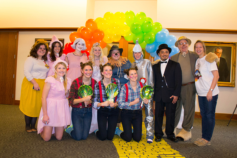 Students, dressed as characters from the Wizard of Oz, pose under a rainbow umbrella. A yellow brick road is seen under them.