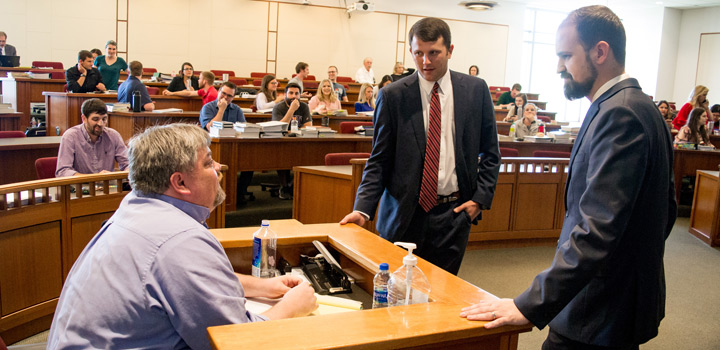 Two students, standing, receive feedback from Coach Robert Little, sitting