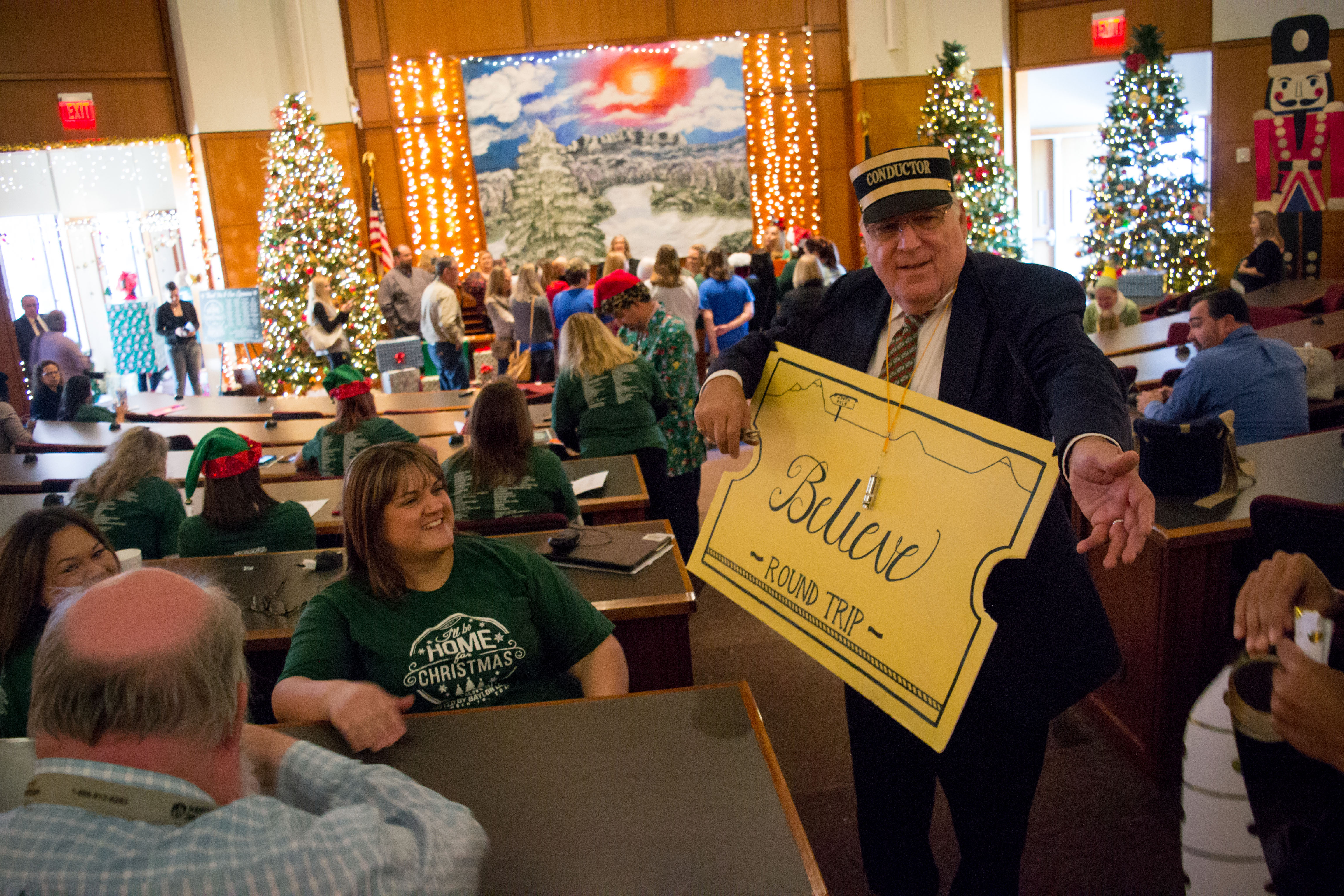 Man dressed as the conductor from 'The Polar Express' entertains families. Several Christmas trees and decorations visible in the background.