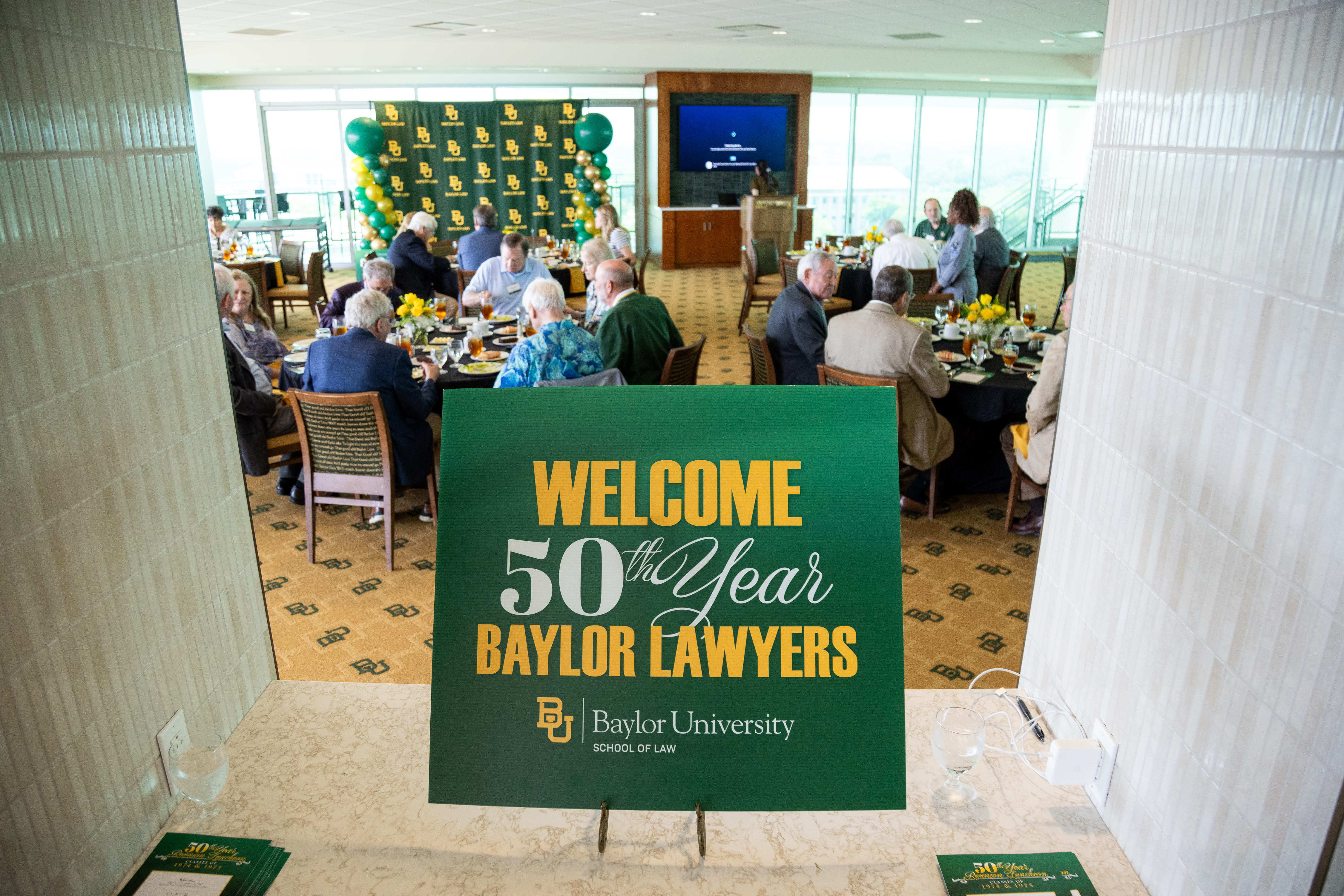 Welcome sign for the 50th reunion in foreground, attendees, seated at tables in the background